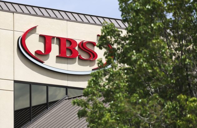 Letreiro vermelho 'JBS' em prédio, com árvore verde em primeiro plano.