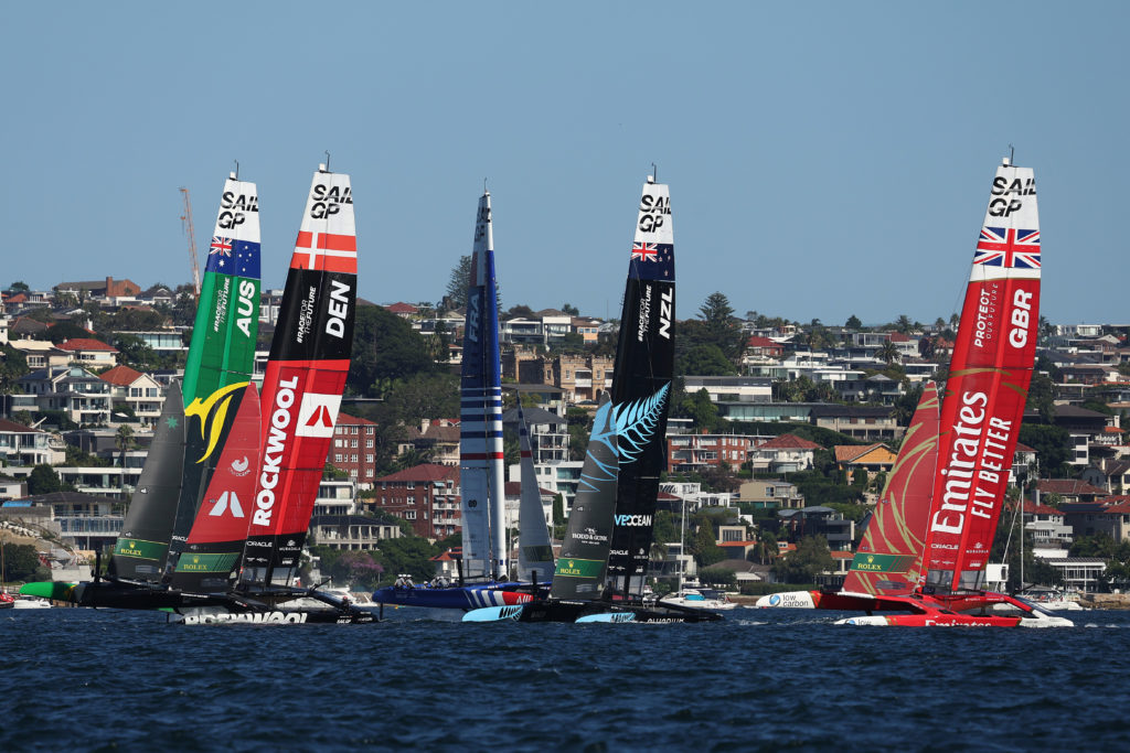Foil-catamarãs de SailGP (AUS, DEN, FRA, NZL, GBR) competem em regata, com casas na costa ao fundo.