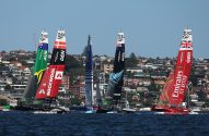 Foil-catamarãs de SailGP (AUS, DEN, FRA, NZL, GBR) competem em regata, com casas na costa ao fundo.
