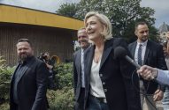 Marine Le Pen sorrindo e falando ao microfone, com homens ao redor e um prédio de madeira com teto amarelo.