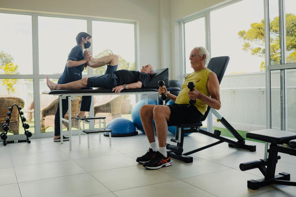 Fisioterapia: terapeuta alonga paciente na maca, enquanto idoso treina com halteres.