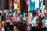 Pessoas andando entre prédios em uma rua iluminada à noite; Seoul, Coreia do Sul