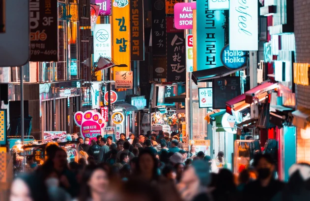Pessoas andando entre prédios em uma rua iluminada à noite; Seoul, Coreia do Sul