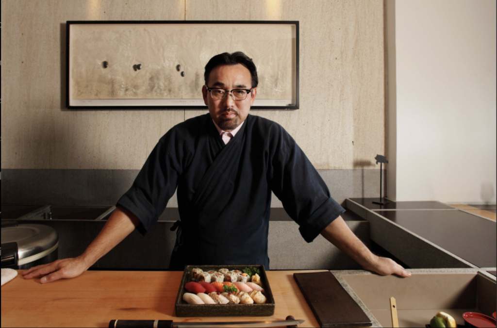 Chef japonês de sushi em uniforme escuro, olhando para a câmera, com bandeja de sushi em balcão.