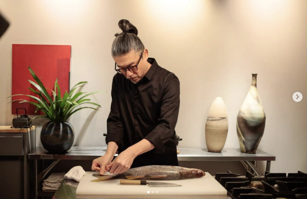 Chef de coque e óculos, camisa escura, fileta um peixe em tábua de corte, com facas.