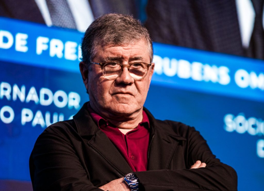 Homem de óculos, camisa vermelha, braços cruzados, em retrato sério. Fundo azul desfocado com texto.