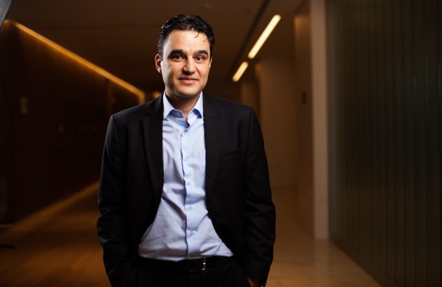 Homem de terno e camisa azul em pé num corredor, sorrindo com as mãos nos bolsos.