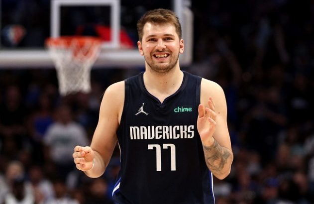 Luka Dončić, jogador de basquete, sorri para a câmera em quadra, vestindo a camisa 77 do Mavericks.