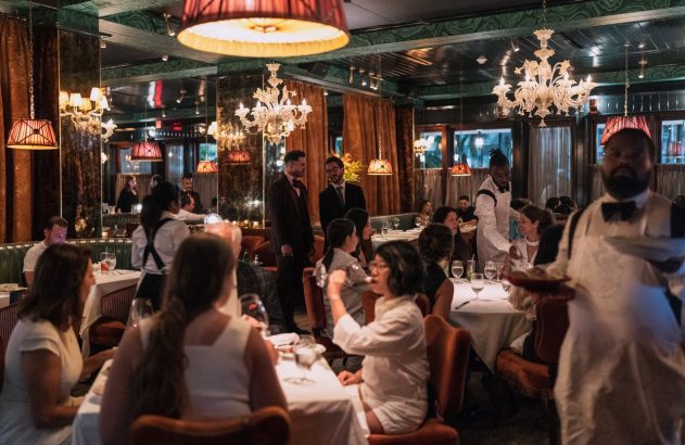 Interior de restaurante elegante com espelhos e teto verde. Clientes jantam em mesas, garçons servem. Ambiente movimentado.
