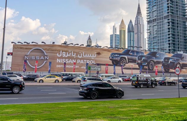 Outdoor da Nissan Patrol com SUVs em rua movimentada de cidade, prédios altos ao fundo.
