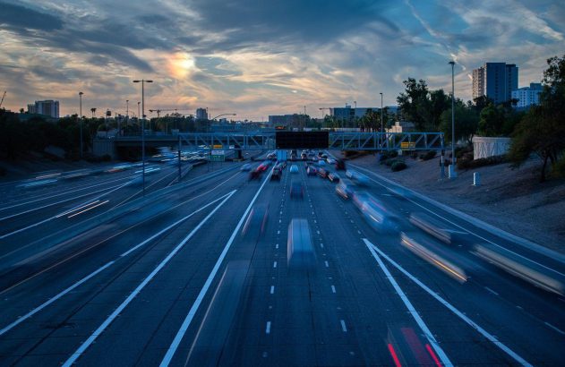 Rodovia movimentada ao entardecer, com carros borrados pelo movimento, sob céu nublado com sol e prédios.