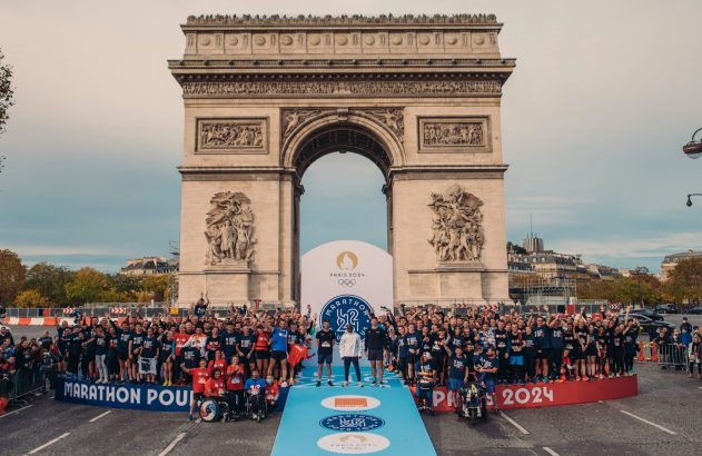 Participantes do Marathon Pour Tous posam em frente ao Arco do Triunfo, com banners Paris 2024.
