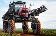 Pulverizador agrícola vermelho em estrada de terra no campo verde. Operador na cabine.