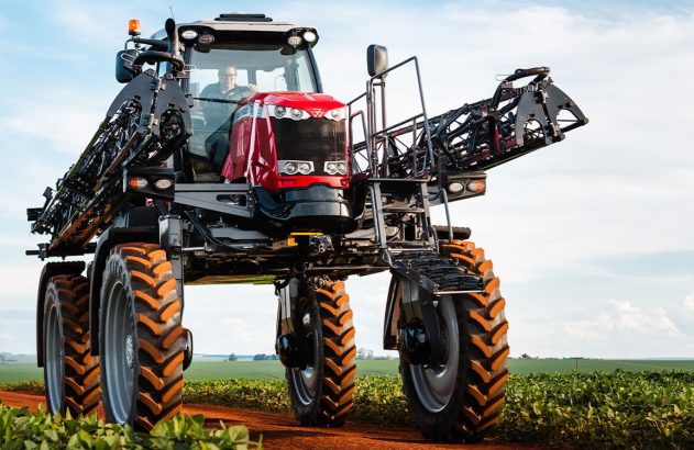 Pulverizador agrícola vermelho em estrada de terra no campo verde. Operador na cabine.