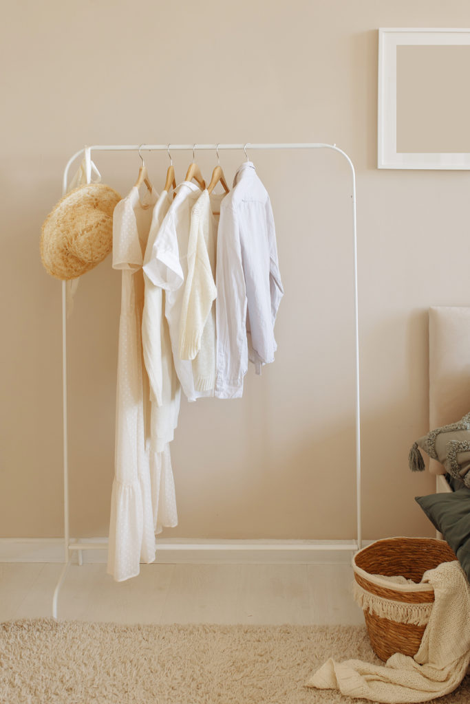 Arara de roupas brancas com peças claras e chapéu em um quarto bege.