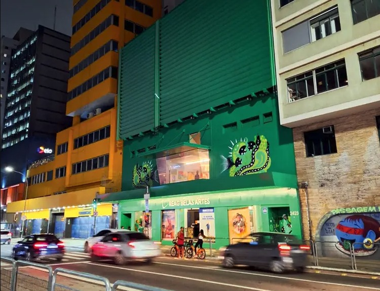 Edifício verde vibrante com arte e painéis à noite, carros em movimento e pessoas em calçada urbana.