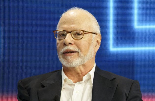 Homem idoso de barba branca e óculos, falando, contra fundo azul e vermelho.