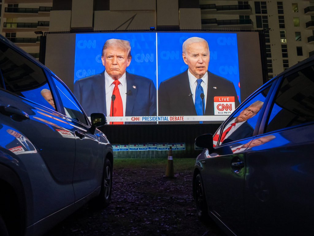 Debate presidencial Trump e Biden em telão, assistido por carros em um drive-in noturno.
