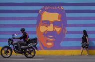 Homem de moto e mulher andando em frente a um mural de rosto com listras azuis e roxas.
