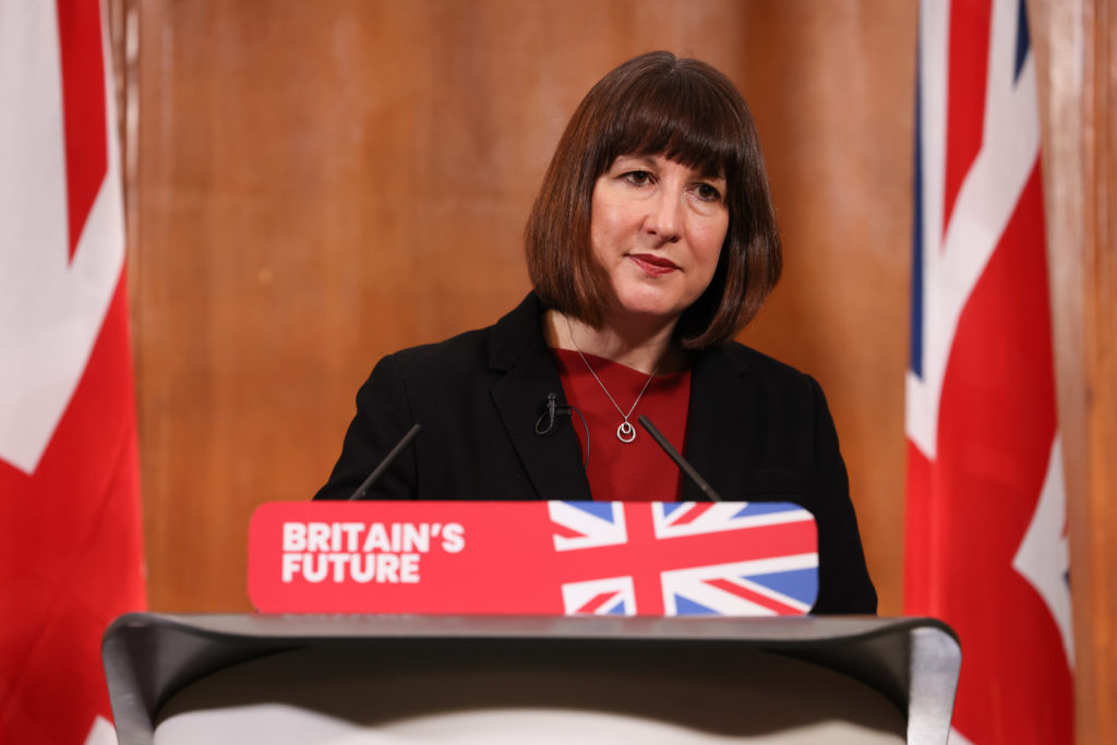 Mulher discursa em pódio com o logo 'BRITAIN'S FUTURE' e bandeiras do Reino Unido ao fundo.