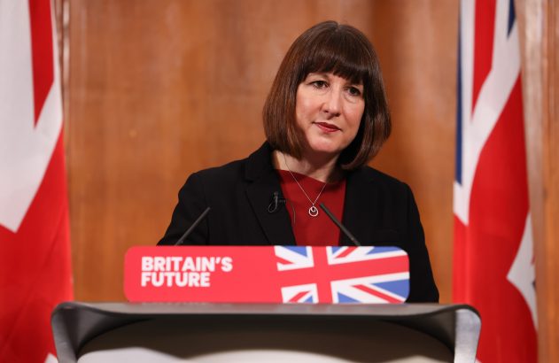 Mulher discursa em pódio com o logo 'BRITAIN'S FUTURE' e bandeiras do Reino Unido ao fundo.