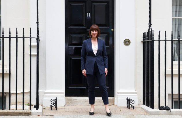 Mulher de terno escuro em pé na porta de um edifício urbano com fachada branca e grades pretas.