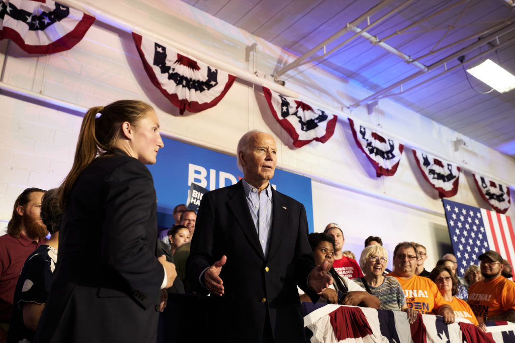 Joe Biden discursa para a plateia em um comício. Bandeiras dos EUA e faixas de campanha ao fundo.