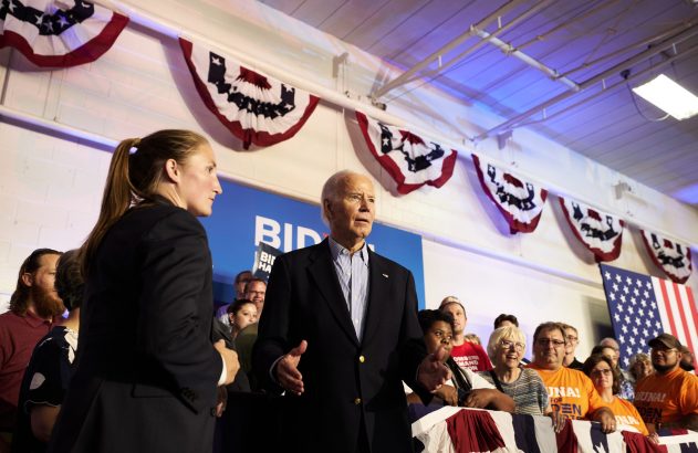 Joe Biden discursa para a plateia em um comício. Bandeiras dos EUA e faixas de campanha ao fundo.