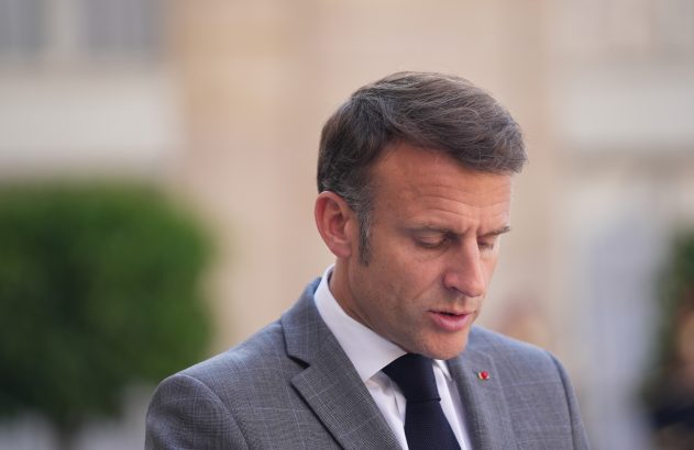 Homem em terno cinza, cabeça inclinada, olhando para baixo, boca levemente aberta. Fundo desfocado.