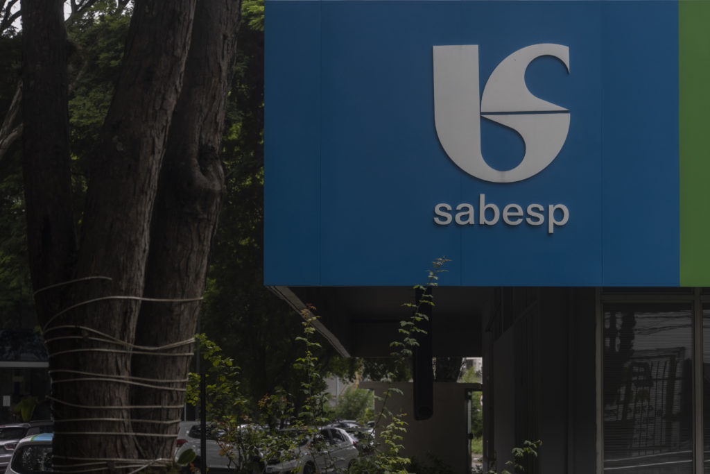 Letreiro azul da Sabesp com logo e nome, em ambiente externo com árvores e parte de edifício.
