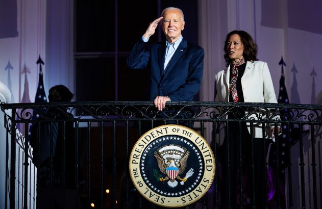 Joe Biden, sorrindo e saudando, e Kamala Harris em uma varanda com o Selo do Presidente dos EUA na grade.