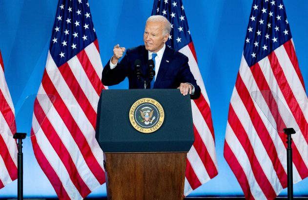 Joe Biden no pódio, cercado por bandeiras dos EUA, com o punho levantado.