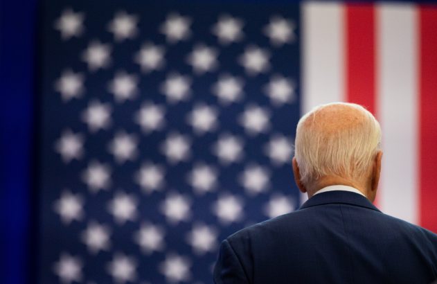 Homem de costas com terno azul e cabelo branco, diante da bandeira dos EUA.