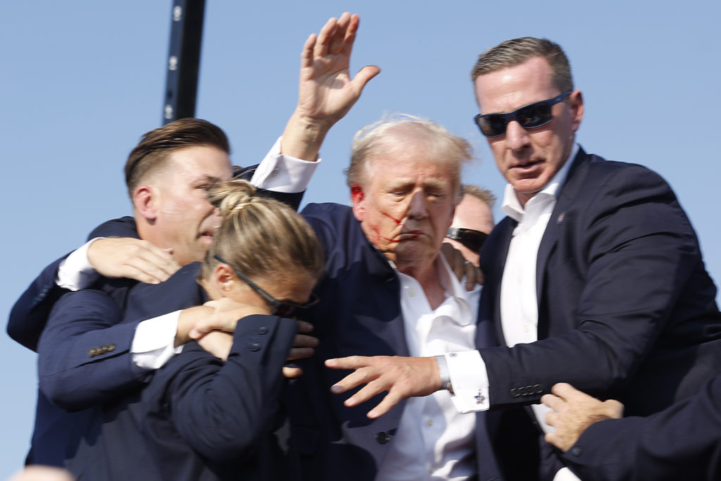 Donald Trump, cercado por seguranças, com mulher de terno e rosto nas mãos. Cena externa.