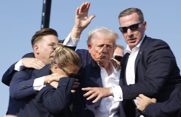 Donald Trump com sangramento no rosto é amparado por vários seguranças. Um braço levantado e céu azul.