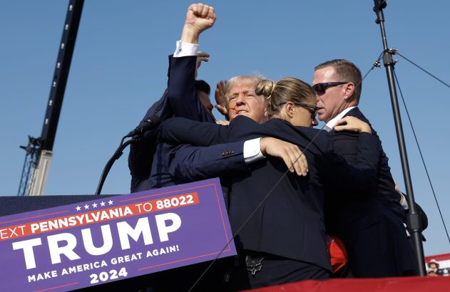 Trump, com punho erguido e ferimento no rosto, é abraçado por equipe em evento político com banner 'Trump 2024'.