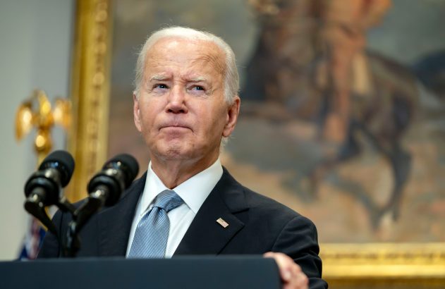 Joe Biden, de cabelos brancos, em um púlpito com microfones, olha para cima. No fundo, moldura dourada e pintura de figura a cavalo.