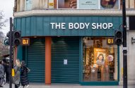 Fachada da The Body Shop, parcialmente com persianas fechadas e vitrine. Pessoas e semáforos com '0.1' e '01' na rua.