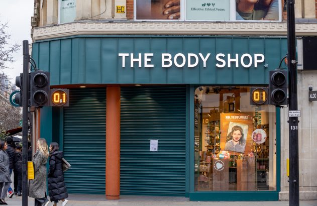 Fachada da The Body Shop, parcialmente com persianas fechadas e vitrine. Pessoas e semáforos com '0.1' e '01' na rua.