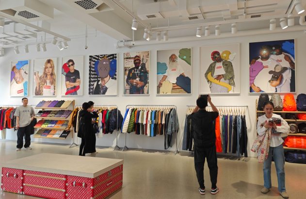 Loja da marca Supreme. Clientes navegam entre roupas expostas em araras e grandes pôsteres com a marca.