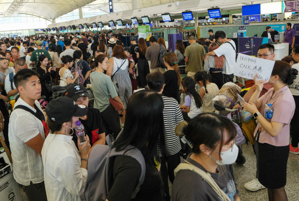 Aeroporto lotado com longas filas de passageiros para check-in. Funcionário exibe placa com informações de voo.