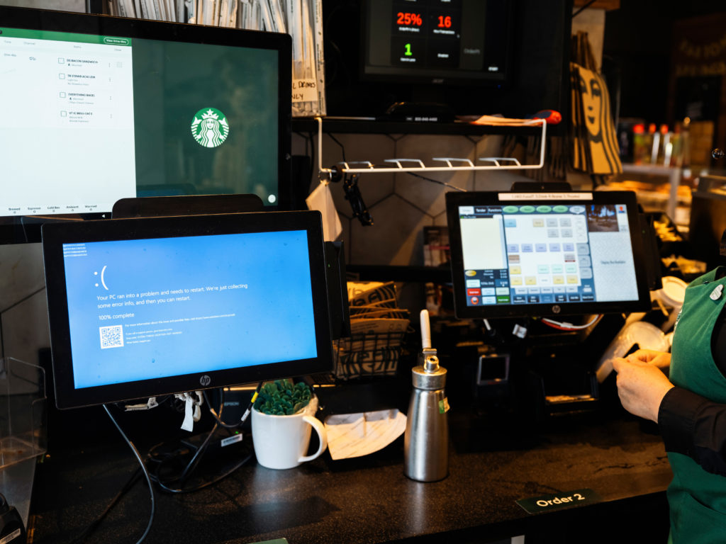 Balcão Starbucks. Uma tela exibe a Tela Azul da Morte, outra o logo. Barista tenta operar os sistemas.