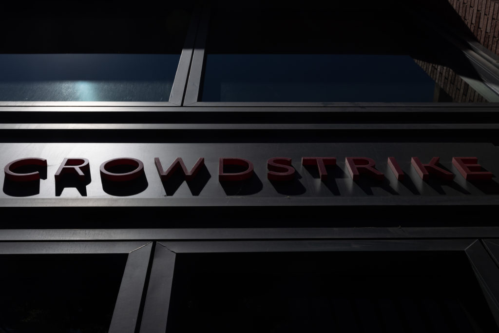 Letreiro vermelho "CROWDSTRIKE" em letras 3D na fachada escura de um edifício, com reflexos em janelas acima.