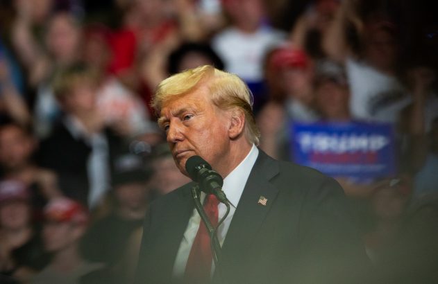 Donald Trump, de terno e gravata, falando a um microfone. Uma multidão embaçada e uma placa "TRUMP" no fundo.