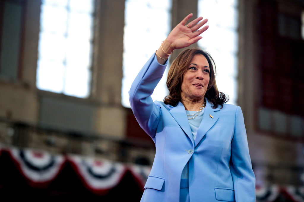 Kamala Harris em terno azul claro acena e sorri em evento.
