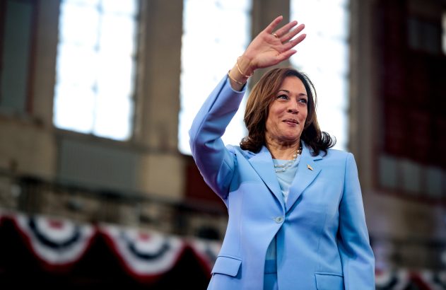 Kamala Harris em terno azul claro acena e sorri em evento.