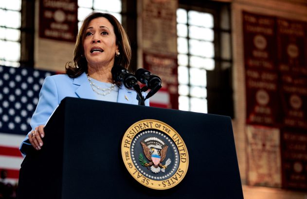 Kamala Harris discursando em pódio com Selo Presidencial e bandeira dos EUA ao fundo.