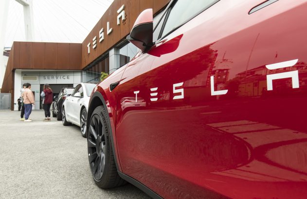 Tesla vermelho em primeiro plano. Edifício Tesla com letreiro 'SERVICE' e pessoas na entrada ao fundo.