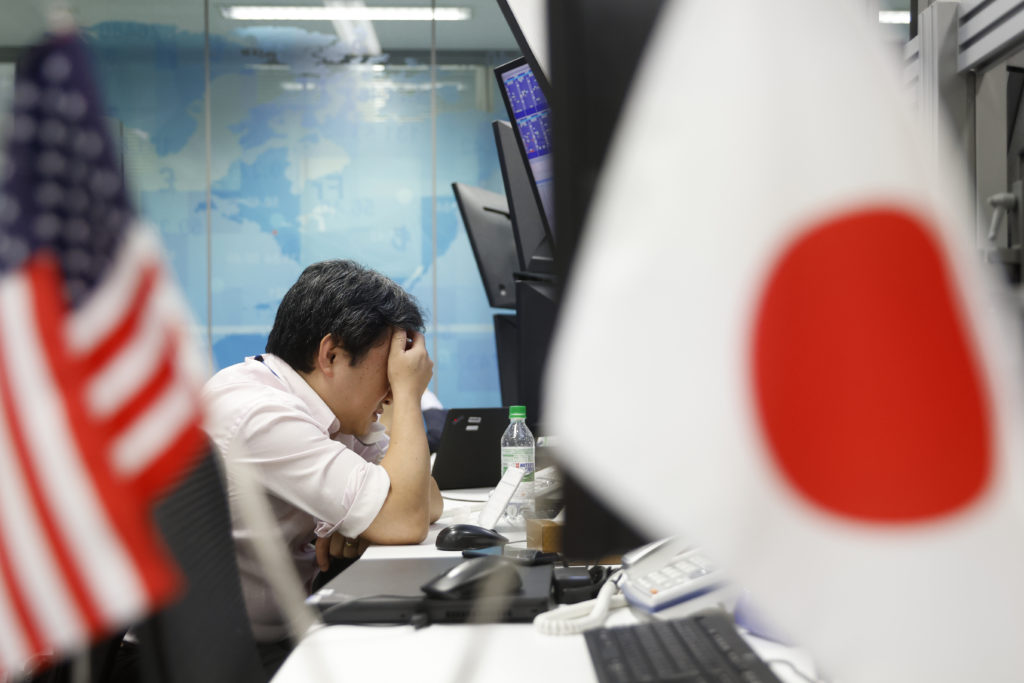 Homem com cabeça nas mãos em escritório, aparentando estresse. Bandeiras dos EUA e Japão.