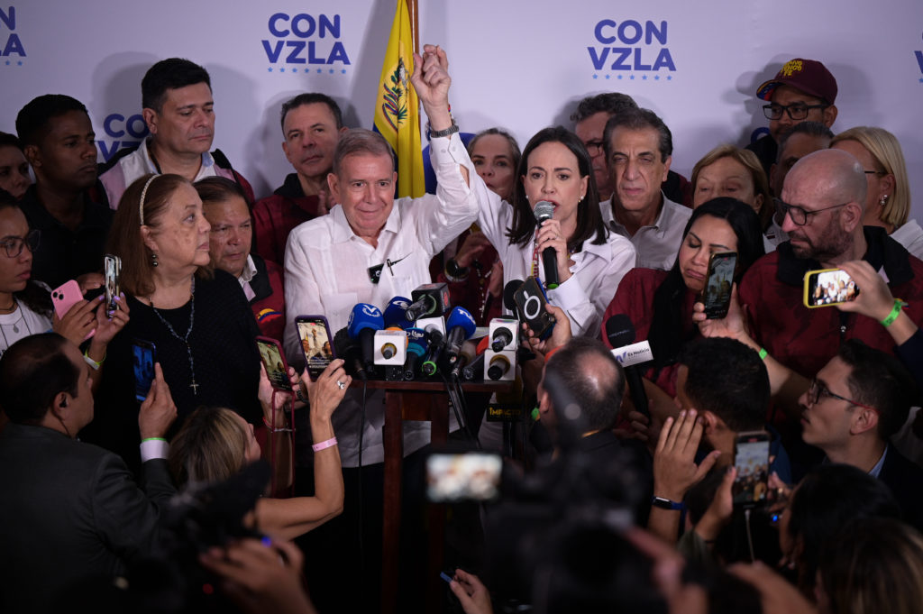 Mulher discursa em microfone, homem com braço erguido ao lado, cercados por pessoas e microfones de imprensa. Banner 'CON VZLA'.